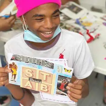 A joyful boy wearing a pink cap and a face mask holds up a collage art project at a table with scattered art supplies.