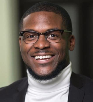 A smiling man with glasses wearing a turtleneck and suit jacket.