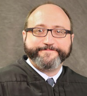 A middle-aged man with a beard, wearing glasses and a judicial robe, smiling at the camera.