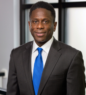 A professional portrait of a middle-aged black man wearing a dark suit and tie, smiling in a modern office setting.