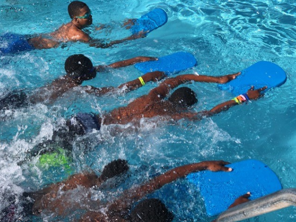 Children swim and play with blue kickboards in a pool, creating splashes.