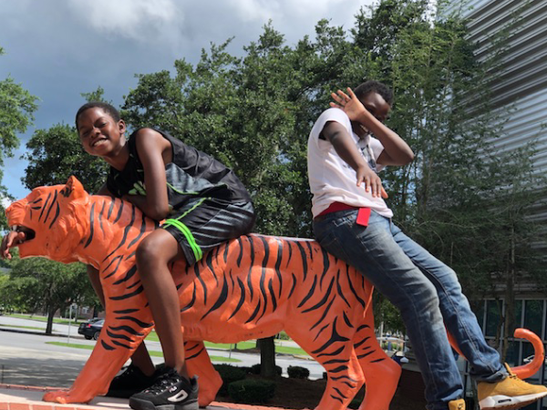 Two boys playing on a brightly painted orange and black tiger sculpture under a sunny sky.