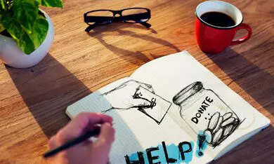 A person sketches a fundraising concept with 'help!' written on paper, accompanied by a coffee cup and glasses on a wooden table.