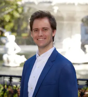 A man in a blue suit and white shirt smiling in front of a fountain and greenery.