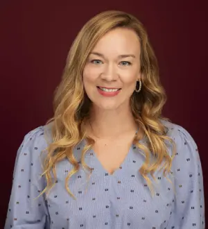 Portrait of a smiling woman with blonde hair wearing a blue blouse, against a maroon background.