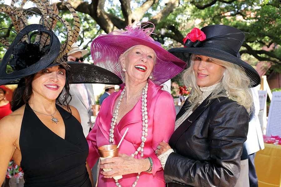 Three elegantly dressed women in extravagant hats enjoy an outdoor social event, smiling as they pose for the photograph.
