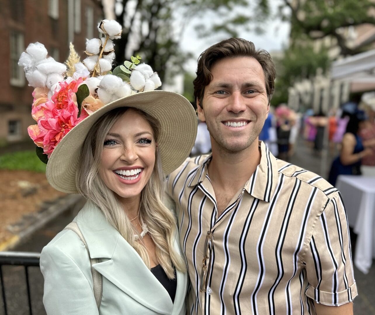 A woman in a decorated hat and a man in a striped shirt smiling together at an outdoor event.