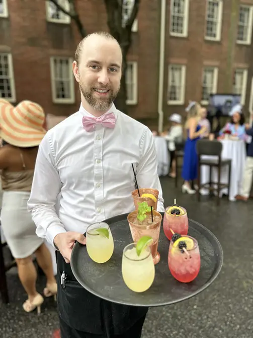 A waiter in a bow tie serving colorful cocktails at an outdoor event, with guests in the background.